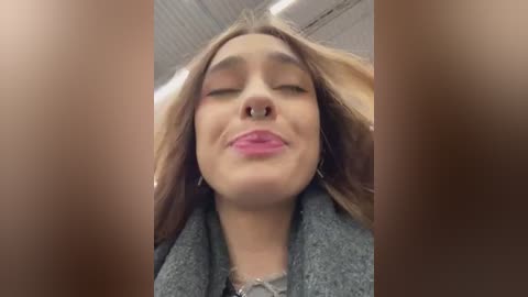 Media: Video of a young woman with long, light brown hair, wearing pink lipstick and a grey sweater, smiling with closed eyes, surrounded by blurred, brown-toned edges.