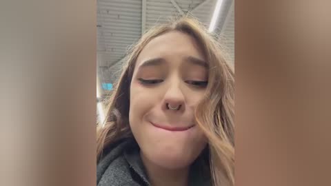 Media: Video of a young woman with fair skin, long wavy blonde hair, and a septum piercing, smiling softly. She wears a dark grey coat. Background shows a modern, industrial ceiling with exposed ductwork.