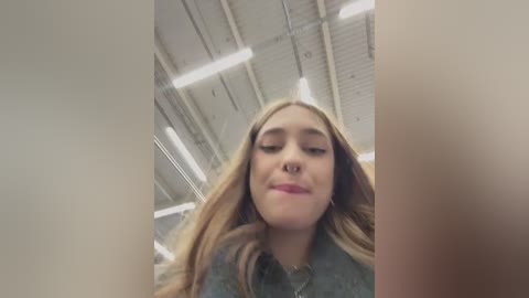 Media: Video of a young woman with long blonde hair, wearing a grey shirt, standing in a warehouse with industrial lighting and exposed ceiling.