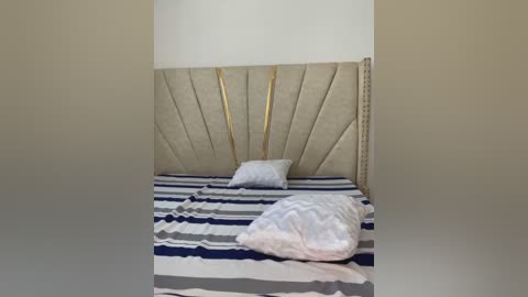 Media: Video of a modern, minimalist bedroom with a beige upholstered headboard, striped bedspread, white pillows, and a neatly folded white blanket on the bed. The room has a plain white wall and no visible furniture or decorations.