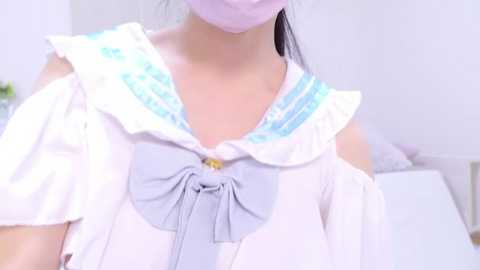 Media: A video of a young woman with East Asian features wearing a pastel sailor uniform, including a large bow tie, and a pink face mask, set in a softly lit room.