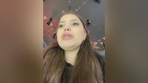 Video of a young woman with long brown hair, fair skin, and pink lipstick, looking upward with a slightly pensive expression, wearing a black top. Background features a dimly lit room with hanging fairy lights.