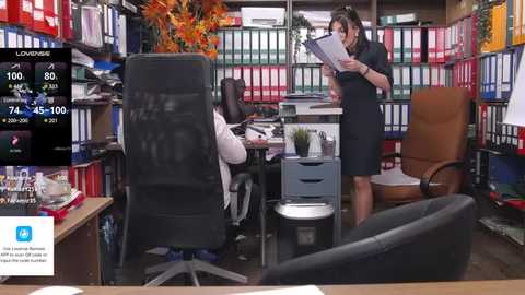 Media: Video of a busy office with two women, one in a skirt suit, one in a blouse, standing and working at desks. Background shows rows of colorful binders and a live news ticker.