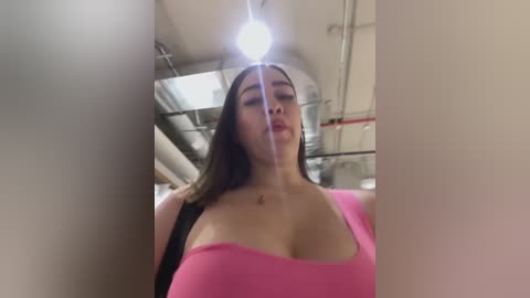 Media: Video of a young woman with long brown hair, wearing a tight pink tank top, inside a brightly lit, industrial space with exposed pipes.