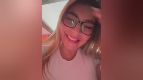 Media: A video of a young woman with long blonde hair, wearing black-rimmed glasses, and a white top, smiling and winking, captured from a low angle, indoors.