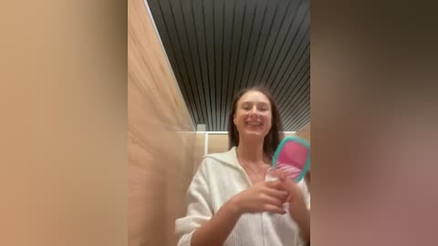 Media: A video of a smiling woman with long hair, holding a pink and blue paddle, standing in a modern bathroom with a wooden wall and black ceiling.