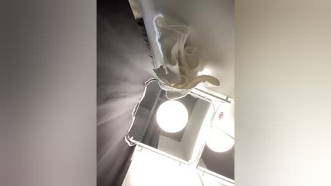 Media: Video of a ceiling fan in a dimly lit room with a white wall, black curtains, and a round light fixture. The fan blades appear motionless, casting a slight shadow.