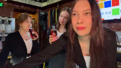 Media: Video of three young women in a bar, one with a large drink, two with smaller drinks. Background shows a TV screen, clothing racks, and a green exit sign.