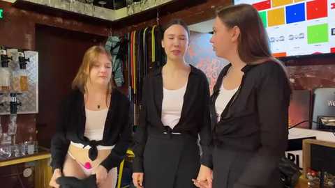 Media: Video of three young women in casual attire, including one wearing a black jacket, standing in a cozy, cluttered room with hanging clothes, colorful posters, and a television.