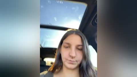 Media: Video of a young woman with long, dark hair, looking down, inside a car with a sunroof open, showing a clear blue sky.