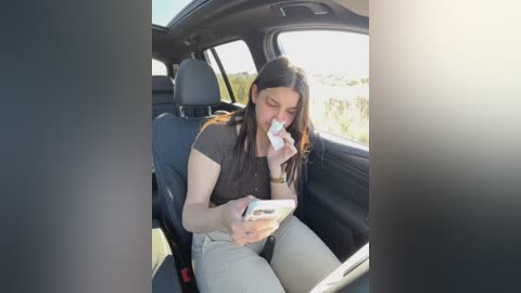 Media: Video of a woman with long brown hair, wearing a black shirt and light gray pants, sitting in a car, reading a newspaper while holding a tissue, in a sunny, open countryside scene.