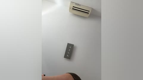 Media: A video of a minimalist, white-walled room featuring a beige air conditioner and a black wall-mounted control panel with three circular buttons. A person's hand, wearing a dark shirt, is visible at the bottom left.