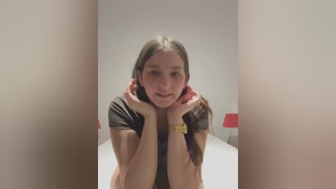 Media: Video of a young woman with long brown hair, wearing a black t-shirt and a yellow watch, leaning on her hands, in a minimalistic, white-walled room with a red lamp in the background.