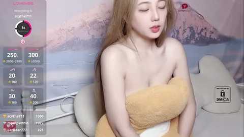 Media: A video of a fair-skinned, petite woman with long blonde hair, wrapped in a beige towel, sitting on a white couch in a room with a pastel wall mural.