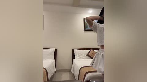 Media: Video of a small, cozy hotel room with two twin beds, white linens, and beige headboards. A man in a white T-shirt stands in the foreground, partially obscuring the view.