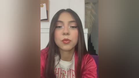 Media: Video of a young Latina girl with long dark hair, wearing a red velvet jacket over a white graphic T-shirt. She has a light skin tone and is indoors, in a dimly lit room with a white wall and a framed photo in the background.