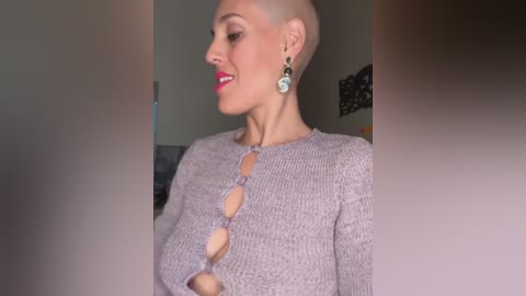 Media: Video of a fair-skinned woman with a shaved head, wearing a gray knitted top with circular cutouts, and turquoise earrings, smiling. Indoor background with blurred furniture.