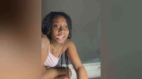 Media: Video of a young Black woman with medium-dark skin and braided hair, wearing a light pink spaghetti strap top, smiling and sticking out her tongue, indoors with a bed and wall in the background.