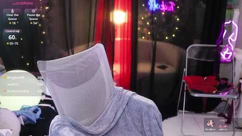 Media: Video of a person in a VR headset with a grey cloth over their head, seated in a dimly lit room with a pink and purple neon sign, a red curtain, and a table with scattered items.