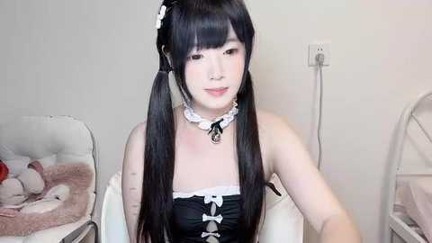 Media: A video of a young Asian woman with long black pigtails, fair skin, and a delicate, petite frame, wearing a black maid outfit and white lace choker, seated in a minimalist bedroom with a white bed and a stuffed animal.