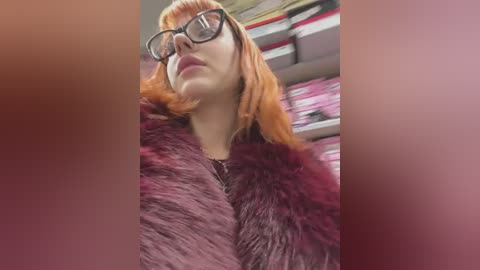 Media: Video of a woman with straight, shoulder-length, orange hair, wearing black-rimmed glasses, a dark fur coat, and red lipstick, standing in a cluttered, pink-shelved room.