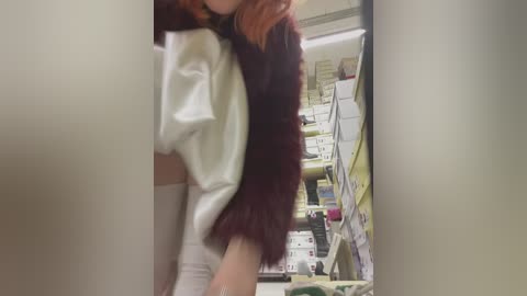 Media: A video of a person with reddish hair, wearing a white top and dark furry jacket, standing in a cluttered, brightly-lit pharmacy aisle filled with various products.