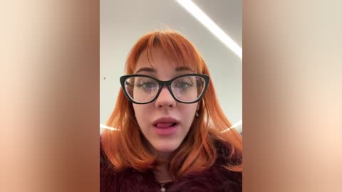 Media: Video of a young woman with fair skin, orange hair, and black-framed glasses, wearing a maroon jacket, captured indoors with a blurred background and bright lighting.