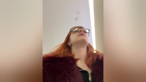 Media: A blurry video of a young woman with fair skin, red hair, and wearing reflective sunglasses, a maroon fur coat, and a black top, standing in a bright, modern hallway.