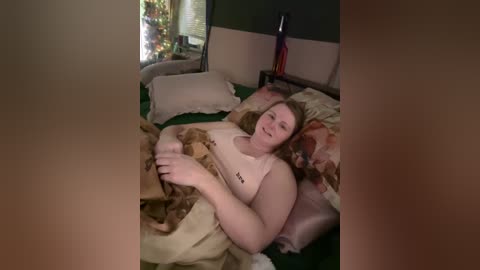 Media: Video of a smiling, overweight, fair-skinned woman with long brown hair, wearing a beige tank top, lying in a messy bed with a green comforter, surrounded by pillows and a nightstand with a candle.