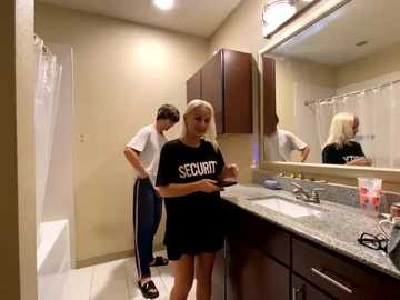 Media: Video of a blonde woman in a black \"SECURITY\" t-shirt, adjusting her pants, in a modern bathroom with beige walls, dark cabinets, and a granite countertop.