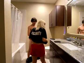 Media: Video of a blonde woman in a black t-shirt and red shorts, standing in a bathroom with a shirtless man, looking at a mirror.