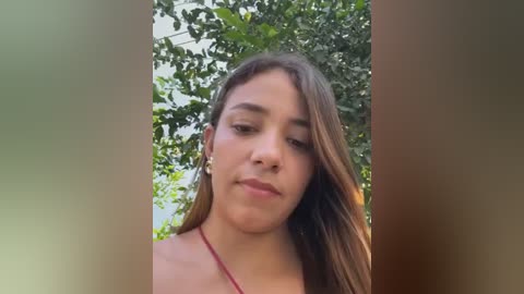 Media: Video of a young Latina woman with light brown skin, long brown hair, and a slim build, wearing a red tank top, standing in a lush, green outdoor setting with tree leaves in focus.