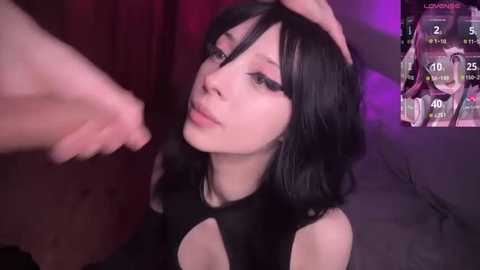 Video of a pale-skinned, slender Asian woman with black hair, wearing a black cutout top, being touched on the head by another person. Background includes a dimly lit room with purple and red lighting.