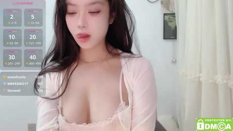 Media: A video of a young Asian woman with long black hair, fair skin, and full lips, wearing a pink lace bra, standing indoors with a calendar and curtains in the background.