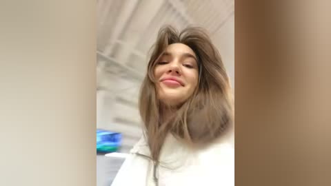 Media: Video of a smiling young woman with shoulder-length, voluminous, light brown hair, wearing a white shirt, standing in a brightly lit room with beige walls and a blue object in the background.