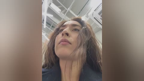 Media: Video of a young woman with medium-length, slightly messy brown hair, wearing a black shirt, captured from a low angle. Background features a modern, industrial-style ceiling with white lights and air conditioning vents.