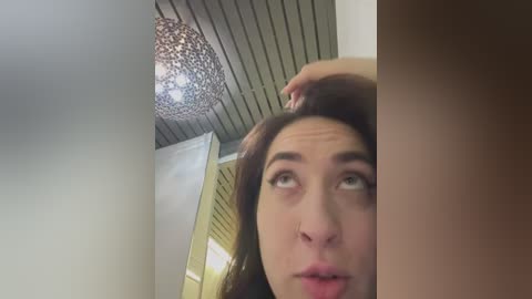 Media: Video of a young woman with light skin and brown hair, looking up with a puzzled expression. The background features a modern interior with a textured ceiling and a chandelier, creating a soft, warm ambiance.