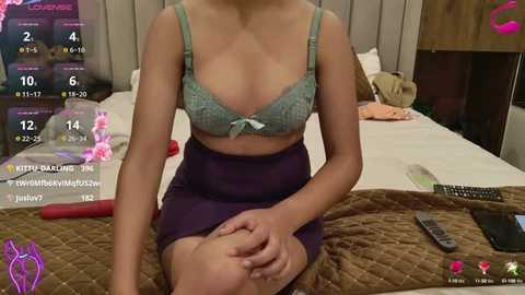 Media: A video of an Asian woman with fair skin, wearing a green lace bra and purple skirt, sitting on a bed in a bedroom, surrounded by various objects, including a TV remote.