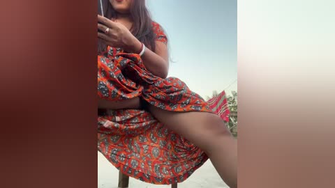 Media: Video of a woman with medium skin tone, wearing a red and orange patterned dress, sitting on a chair outdoors. The background includes a blue sky and a blurred red and white patterned object.