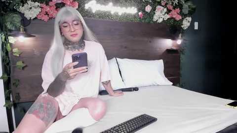 Media: Video of a fair-skinned woman with long platinum hair, tattoos, and piercings, wearing a white nightgown, sitting on a bed with floral decorations and a keyboard on a table.