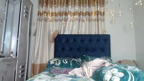 Media: Video of a modern bedroom with a dark blue tufted headboard, teal and gold-patterned bedspread, and a metallic-framed mirror on the left. Hanging from the ceiling are string lights, and beige curtains with gold and white stripes cover the window.