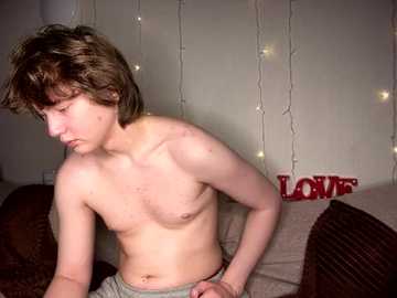 Media: Video of a shirtless, fair-skinned teenage boy with tousled brown hair, wearing grey sweatpants, sitting on a couch. Background features string lights and a \"LOVE\" sign.