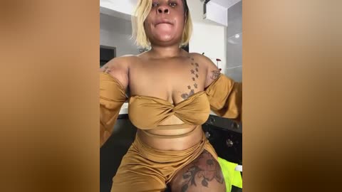 Media: A video of a plus-size Black woman with a short, blonde bob haircut, wearing a mustard-yellow off-shoulder crop top and matching high-waisted shorts, displaying tattoos on her arms and thigh. The background shows a modern, minimalist interior.