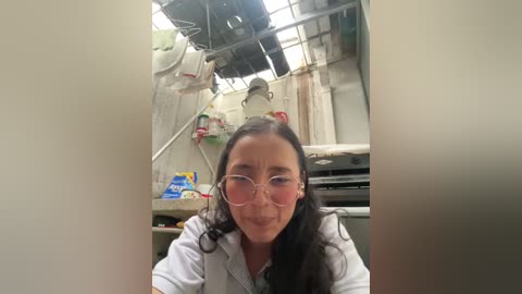 Media: A video shows a woman with long black hair and glasses, wearing a white lab coat, smiling in a cluttered kitchen with hanging pots and a messy countertop.