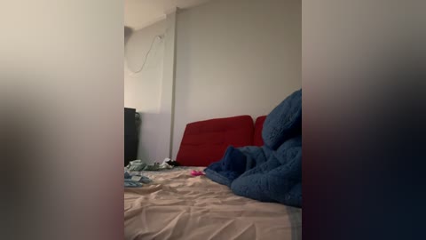 Media: A dimly lit video of a messy bedroom with a beige bedspread, blue blanket, red pillow, and scattered clothes. The walls are plain white, and a black speaker stands against the left wall.