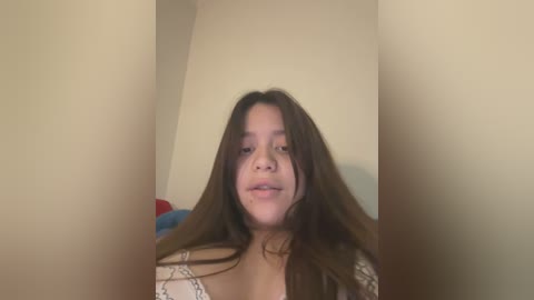 Media: Video of a young Hispanic woman with long, dark brown hair and light skin, wearing a white shirt, standing in a dimly lit room with beige walls.