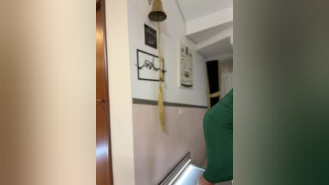 Media: A video of a narrow, beige-tiled hallway with a green wall, featuring a yellow wall-mounted lamp, framed art, and a person in a green shirt partially visible.