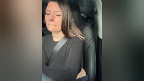 A video of a young woman with long brown hair, wearing a gray sweater, sitting in a car with a seatbelt on, eyes closed, seemingly asleep.