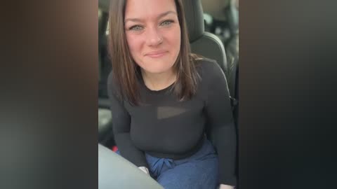 Media: A video of a smiling young woman with light skin and shoulder-length brown hair, wearing a black long-sleeve top and blue jeans, sitting in a car.