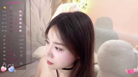 Media: Video of an Asian woman with long, straight black hair, fair skin, and a black choker, looking contemplative. She's in a cozy, pastel-colored room with a bed and a soft, white blanket. The image includes a screen overlay with camera settings and a pink bunny icon.